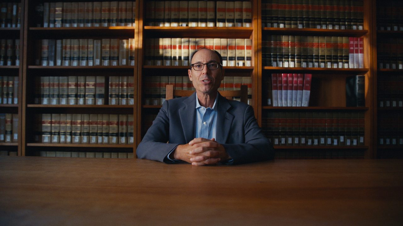 Adam Winkler, in a navy jacket, sits with his hands folded at a long wooden table in a law library lined with books.