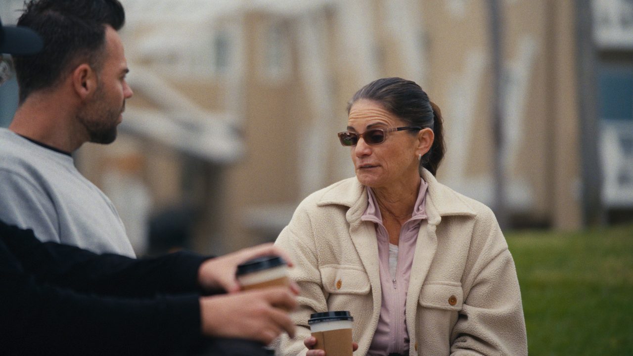 Autumn Bignami, in sunglasses and a cream jacket, holds a coffee and talks with Tommy Jackett on a park lawn.