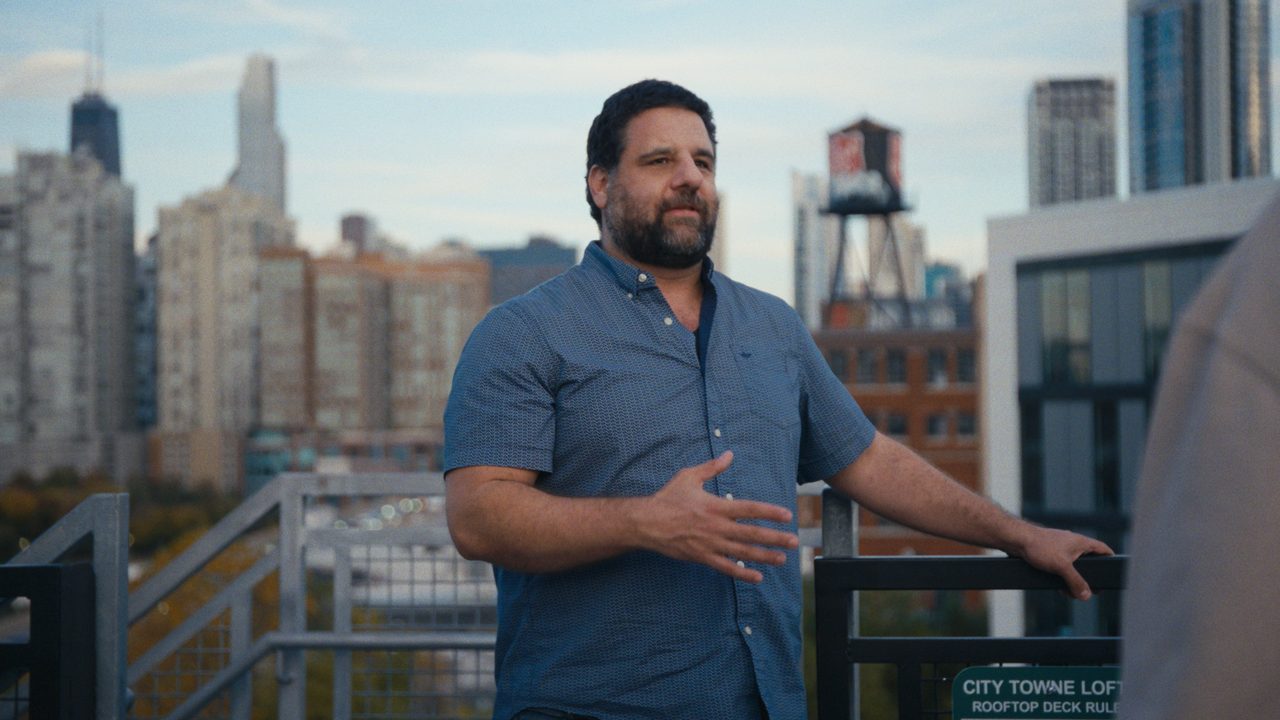 A bearded man in a blue short-sleeved shirt gestures as he speaks on a rooftop deck, with a water tower and high-rises behind him.
