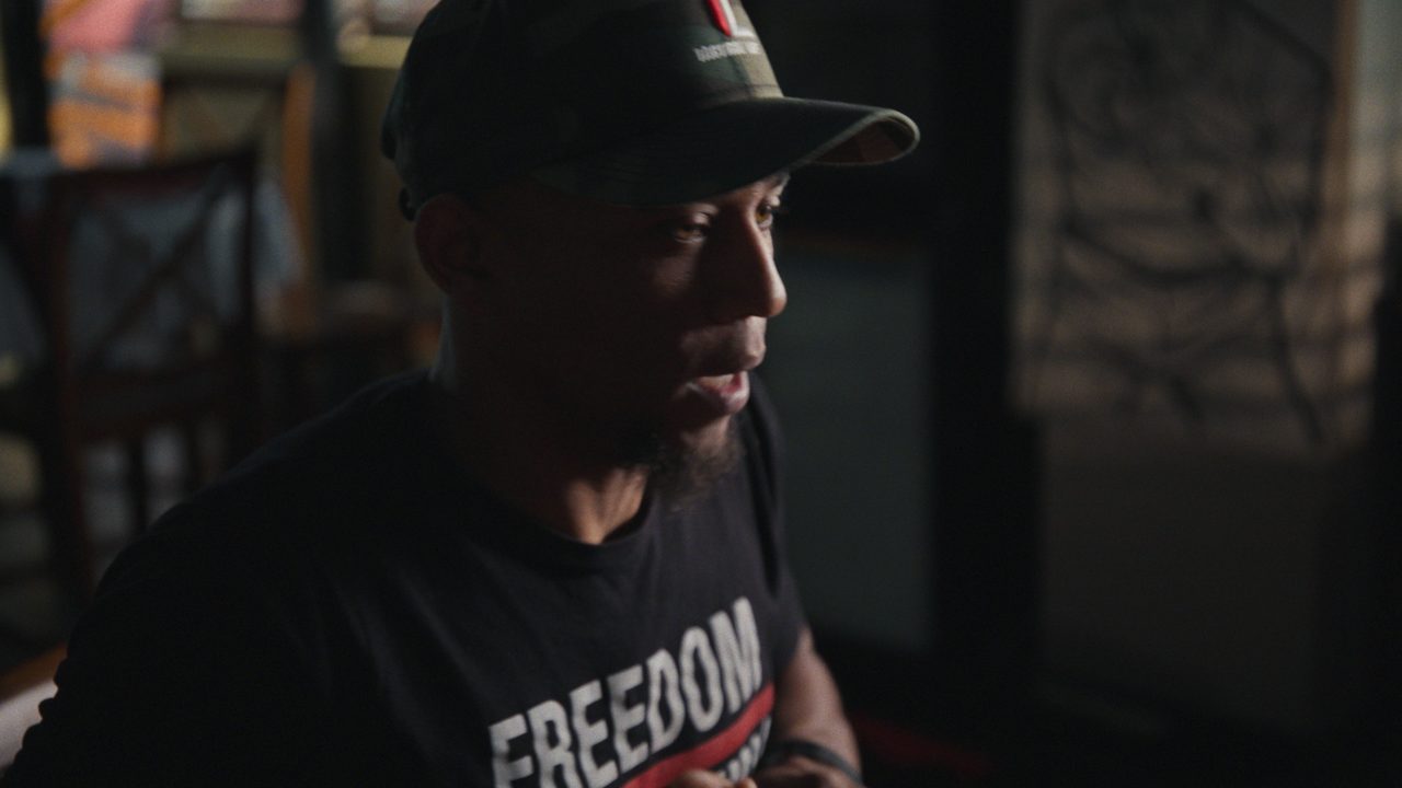 A man in a camouflage cap and a black T-shirt that reads FREEDOM speaks in a dim room.