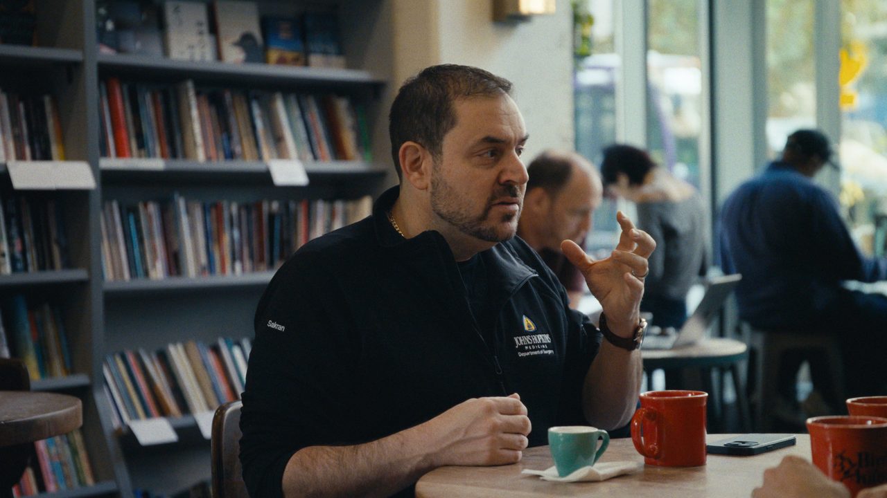 Dr Joseph Sakran gestures as he talks at a café table with coffee cups, bookshelves behind him.