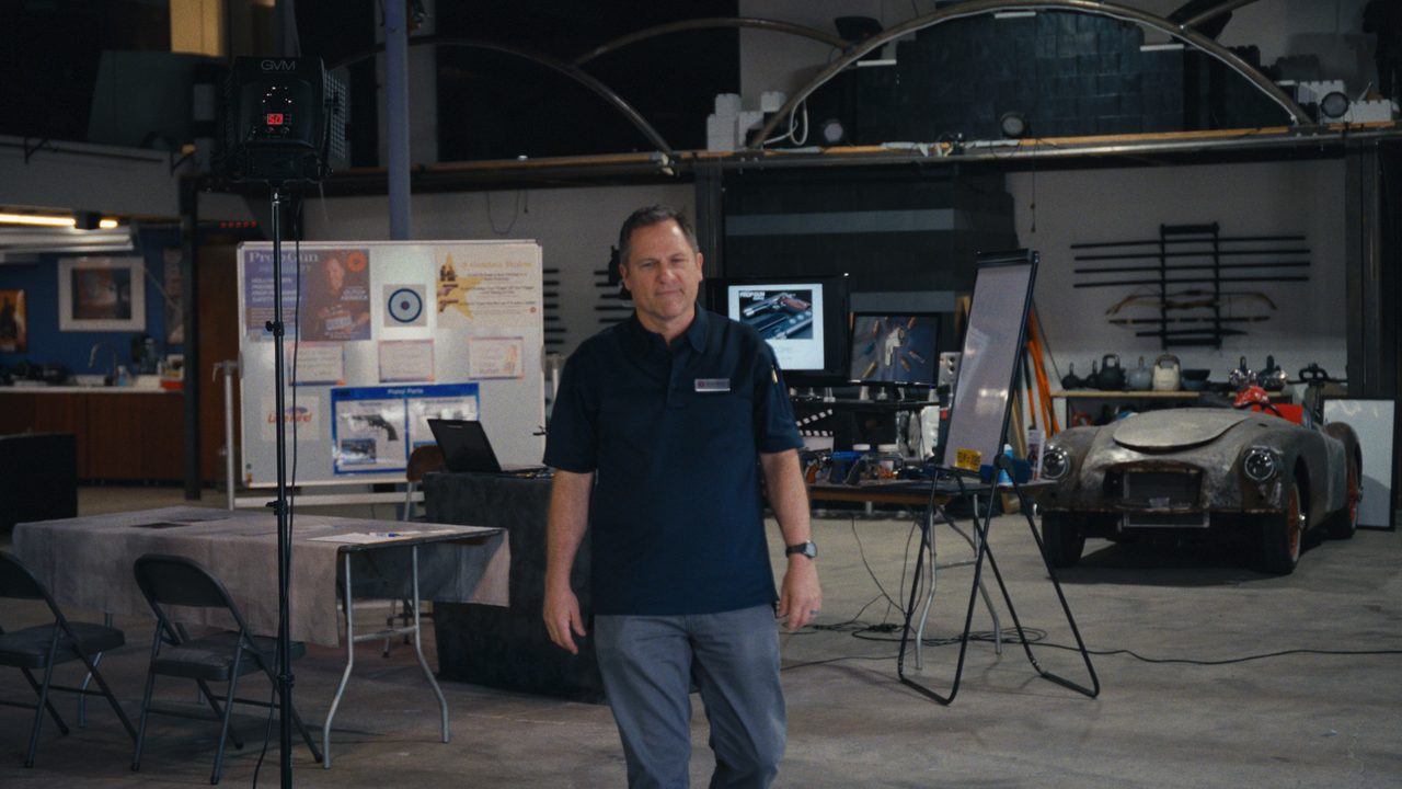 Dutch Merrick stands in a large warehouse among film props, display boards and a vintage car.