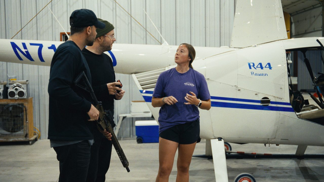 A young woman in a purple T-shirt talks to two men in black beside a white helicopter in a hangar.