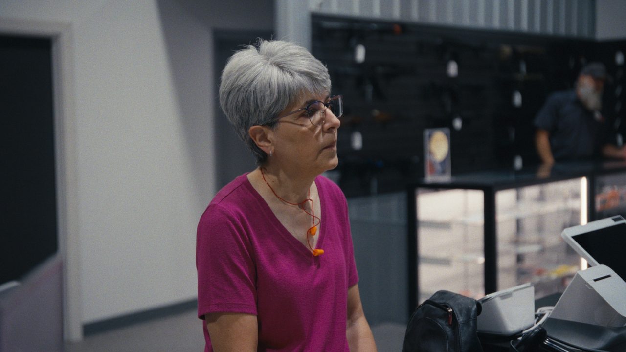 Gina McComb, with short grey hair and glasses, talks inside a gun shop with display cases behind her.