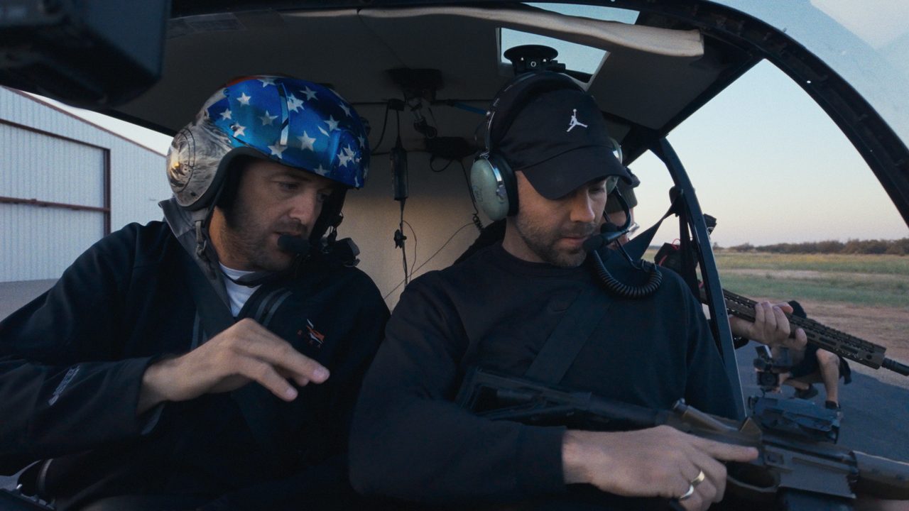 Dustin Johnson in a stars-and-stripes flight helmet sits beside Tommy Jackett, in a headset, inside a helicopter cabin.