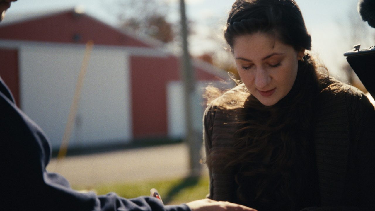 A young woman with dark braided hair looks down as she talks, red and white barns out of focus in the sun behind her.