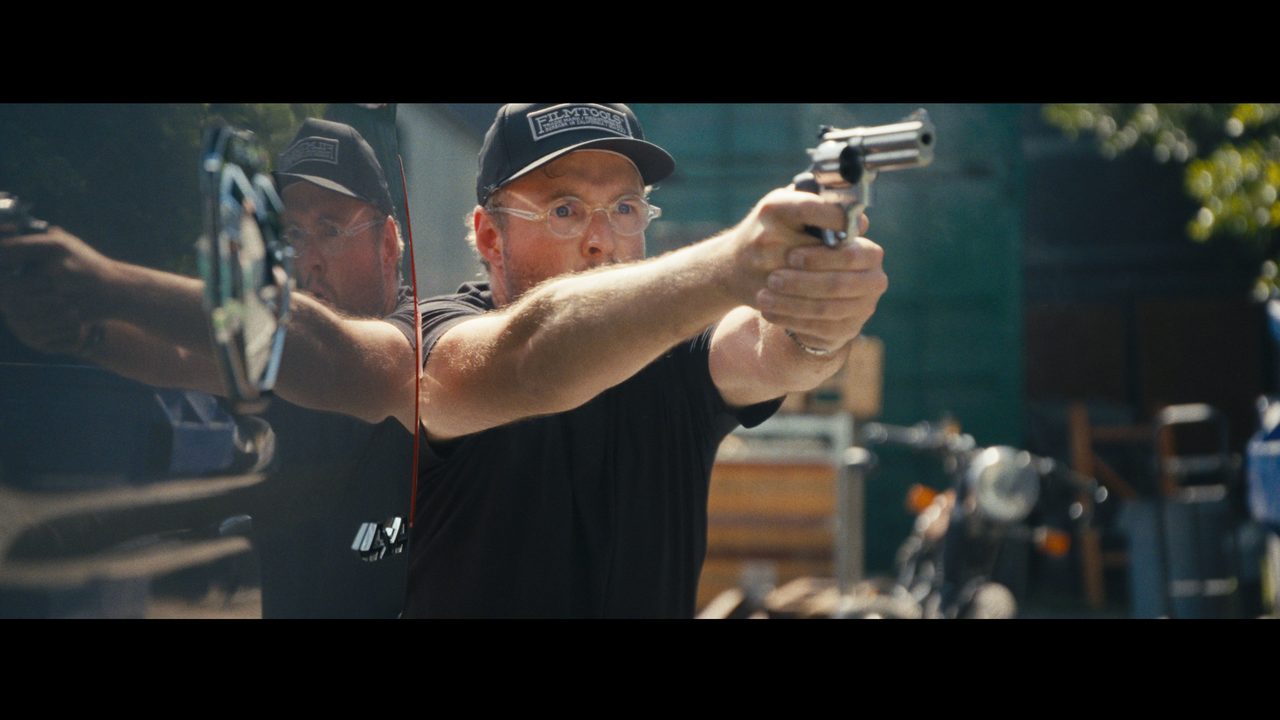 Josh Janssen, in a cap and glasses, aims a revolver with both hands outdoors beside shipping containers.