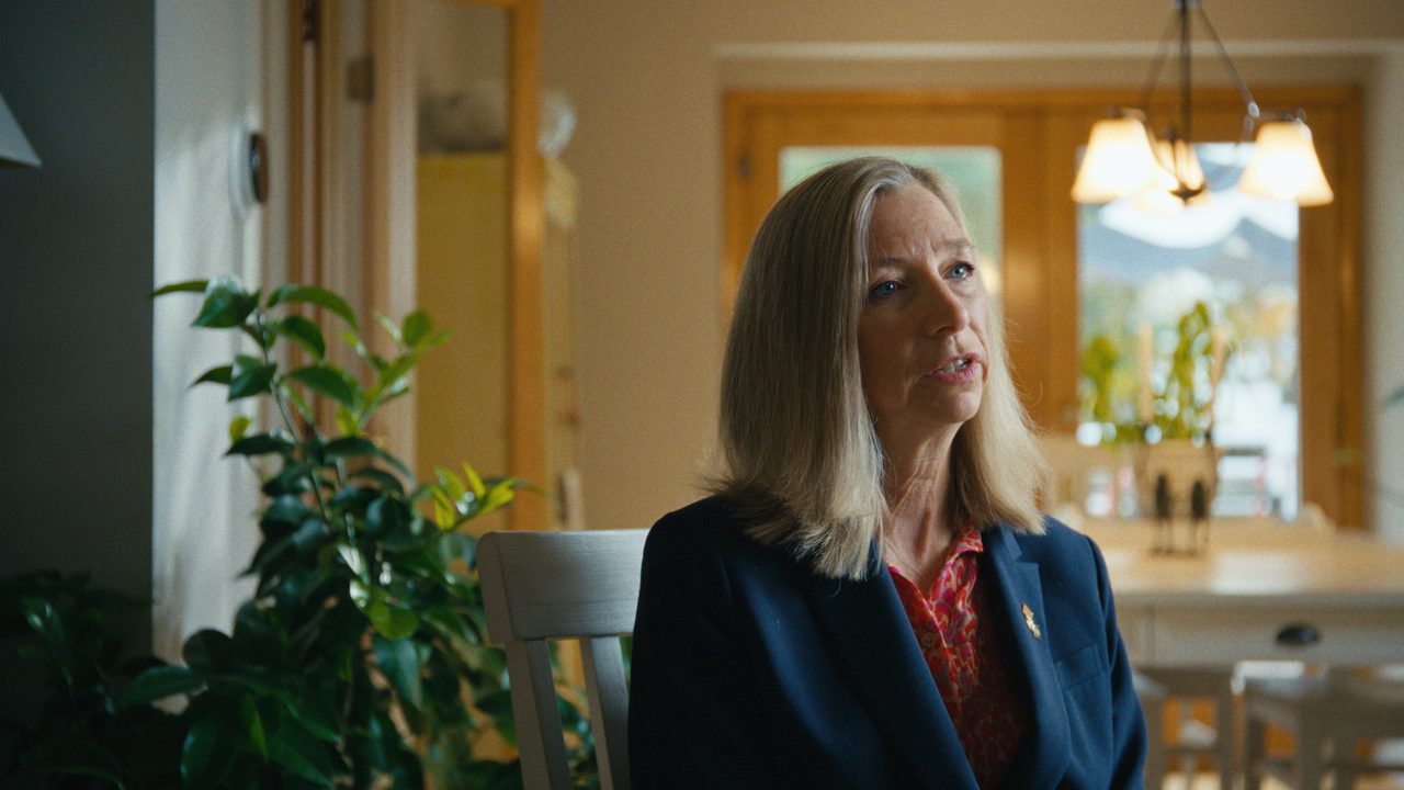 Katherine Schweit, in a navy blazer, listens in a bright room with plants and a chandelier behind her.