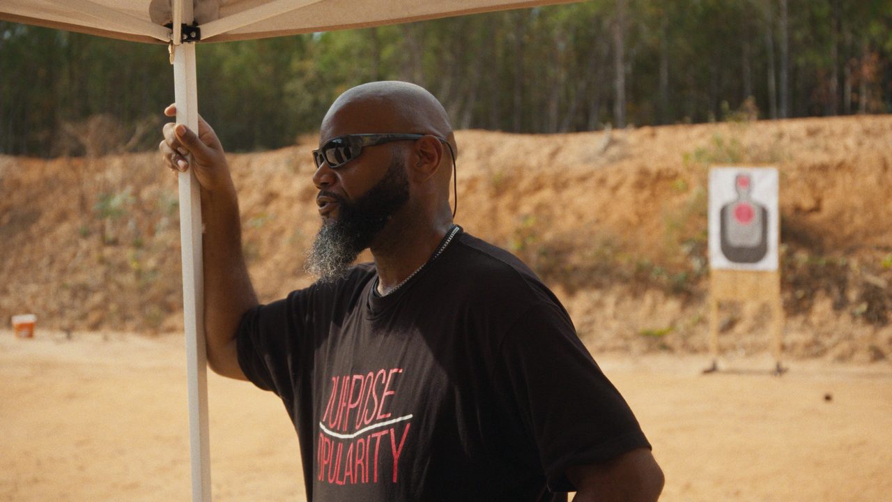 Kevin Dixie, in sunglasses and a 'Purpose over popularity' T-shirt, leans on a tent pole at an outdoor range.