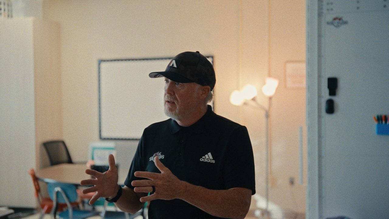 Kevin Thomas, in a black cap and polo shirt, gestures as he explains, inside a classroom with a whiteboard.
