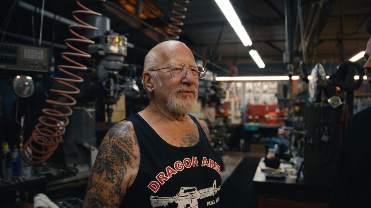 Mel Bernstein, white-bearded with tattooed arms, smiles in his workshop surrounded by machinery.