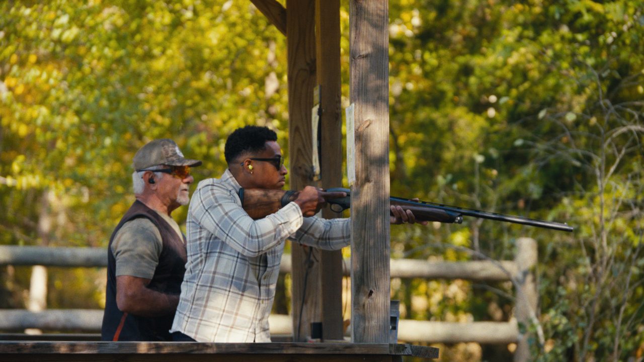 Mychael Waller aims a shotgun from a wooden shooting stand while Joe Calabrace watches beside him.