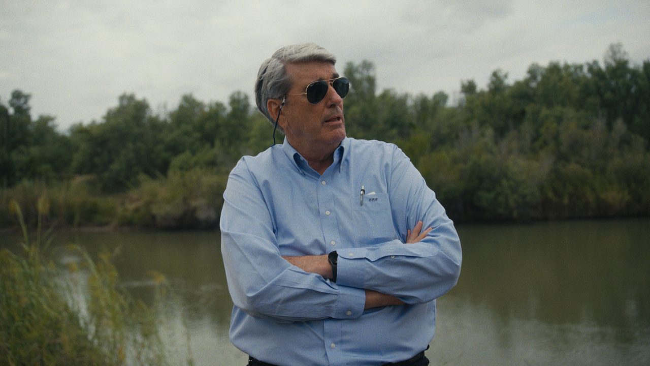 Othal Brand Jr., in sunglasses and a pale blue shirt, stands with his arms folded beside a river lined with trees.