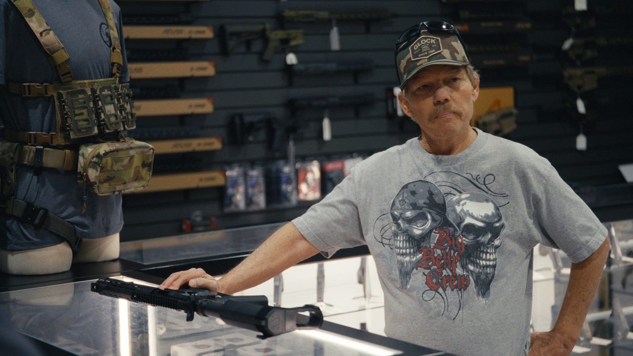 Raymond Erickson, in a camouflage cap and grey T-shirt, leans on a gun-shop counter with a rifle resting under his hand.