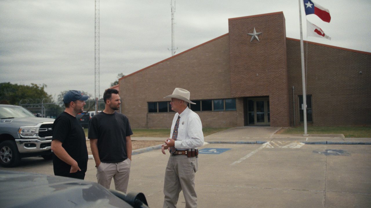 A sheriff in a white shirt, tie and cream cowboy hat talks with two men in black T-shirts in the car park of a brick county building, a Texas flag flying behind.
