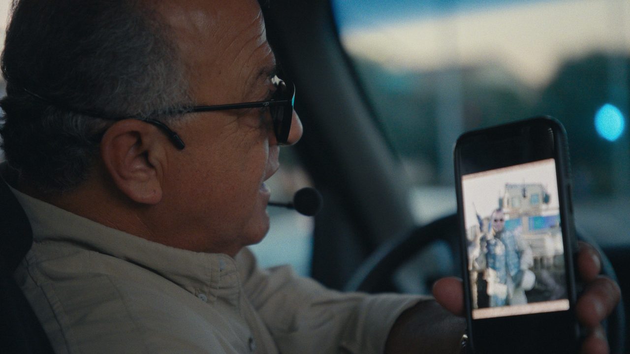 Sammy Dorian, wearing a headset microphone at the wheel of his tour bus, holds up a phone showing a photo of himself in Iraq.