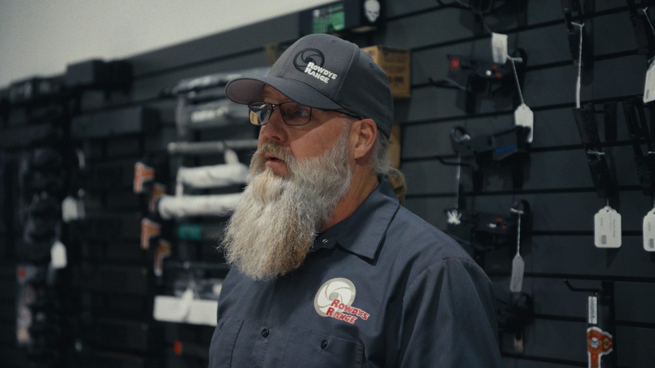 A man with a long grey beard, glasses and a Rowdy’s Range cap and work shirt stands in front of a wall of rifles and handguns hung on display hooks.