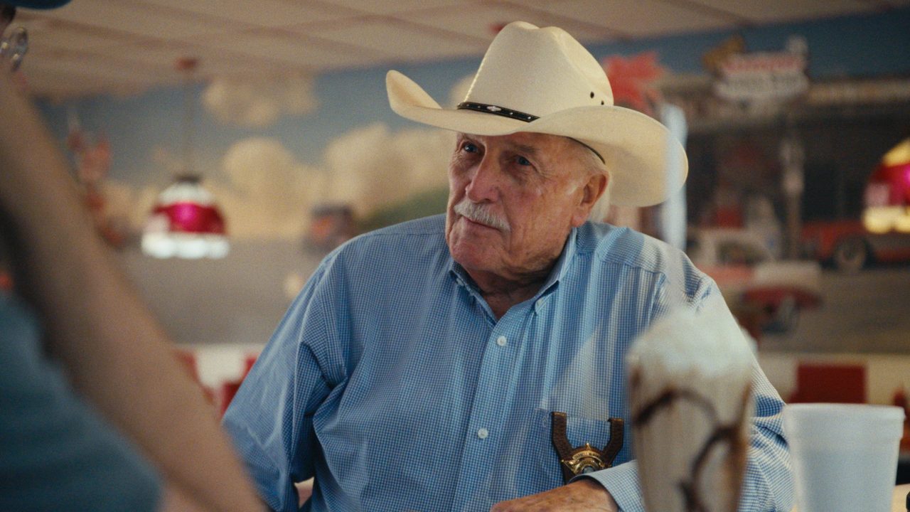 Sheriff Parnell McNamara, in a white cowboy hat and blue shirt, sits in a retro diner booth beside a milkshake.