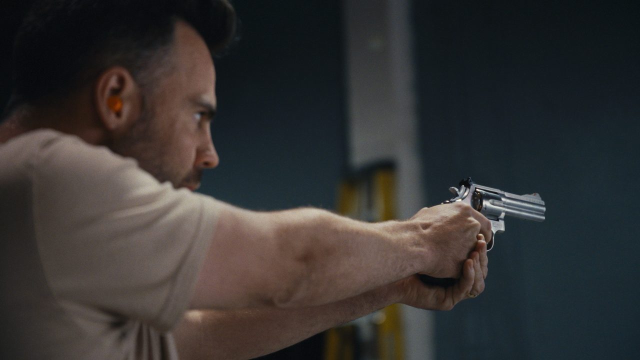 Tommy Jackett, in ear plugs, aims a silver prop revolver with both hands in a dim warehouse.
