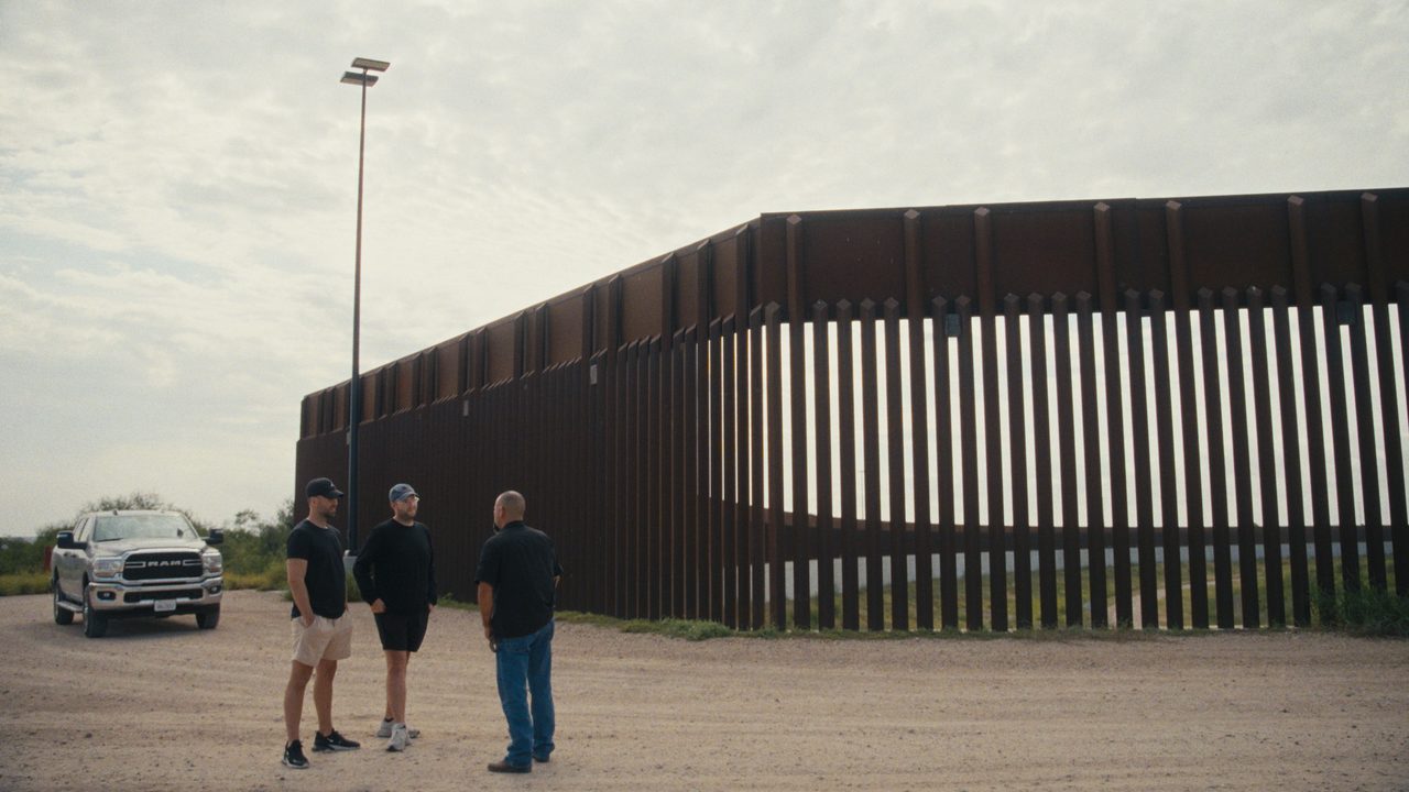 Three small figures stand on gravel beside a tall steel border wall at dusk, with a pickup truck parked nearby.